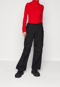 Donna che indossa un maglione fitted rosso con collo alto, pantaloni larghi neri e scarpe nero con zeppa, in piedi contro uno sfondo neutro.