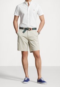 Man i vit kortärmad skjorta med knappar, beige shorts med randig bälte, blå sneakers och armbandsur, med händerna i fickorna, stående.