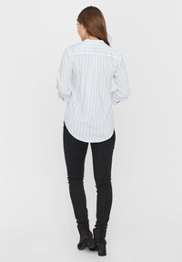 Žena stojící zády, oblečená v světle modré a bílé pruhované košili, černých skinny kalhotách a černých kotníkových botách na bílém pozadí.