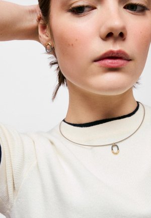Mujer que lleva un suéter crema con detalles negros, pendientes ovalados dorados y plateados, y un colgante a juego en un collar choker fino de plata.