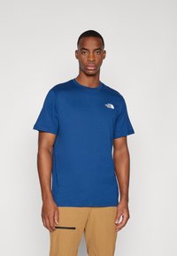 Zila kokvilnas t-krekls ar īsām piedurknēm un apaļu kaklu, ar baltu logotipu kreisajā krūtī. Gluda tekstūra, brīva piegriezuma.