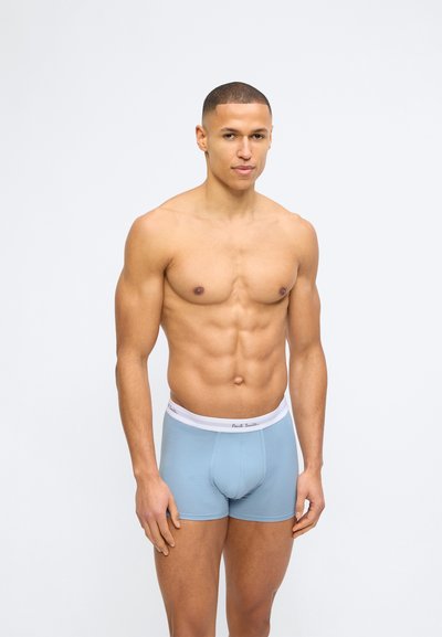 Μυώδης άνδρας στέκεται φορώντας ανοιχτό μπλε ανδρικά εσώρουχα boxer με λευκή ζώνη μέσης, σε απλό λευκό φόντο.