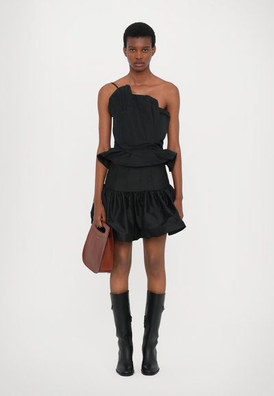 Top asimétrico negro con pliegues, falda negra de capas con volantes, botas negras hasta la rodilla y un bolso marrón. Textura de tela suave.