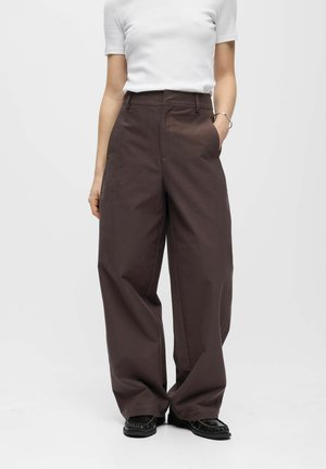 Personne portant un pantalon marron taille haute à jambes larges, un haut blanc ajusté à manches courtes, des chaussures noires et un bracelet argenté, debout devant un fond uni.