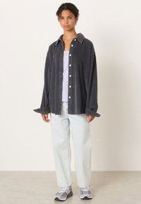 Mlada oseba, oblečena v preveliko temno sivo žametno srajco, svetlo sivi teniški top, svetlo modre široke kavbojke in sive superge, stoji notri.