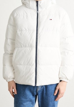 Persoon draagt een witte doorgestikte gewatteerde jas met capuchon en een zichtbare marineblauwe ritssluiting, gecombineerd met blauwe spijkerbroek.