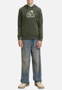 Olijfgroene hoodie met een grafisch ontwerp van een berg en de zon, gecombineerd met losse blauwe jeans. Het model staat op een neutrale achtergrond en toont de outfit.