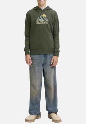 Un sweat à capuche vert olive avec un graphique de montagne et de soleil, associé à un jean bleu ample. Le modèle se tient sur un fond neutre, mettant en valeur la tenue.