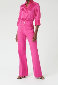 Blouse en satin rose fluo avec un col mandarin et des manches trois-quarts, assortie à un pantalon flared taille haute rose fluo et des sandales à talons noires.