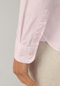 Light pink long-sleeve shirt with a buttoned cuff, featuring a smooth texture, subtle stitching, and a slightly curved hem.