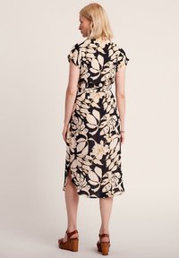Robe noire avec un motif floral en crème et or. Mi-longue avec des manches courtes, une taille nouée et des fentes sur les côtés. Sandales à talons compensés brunes.