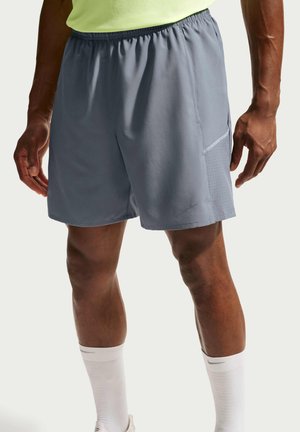 Atleta con pantaloncini sportivi grigi, calzini bianchi e una maglia giallo neon, in piedi su uno sfondo chiaro e uniforme.