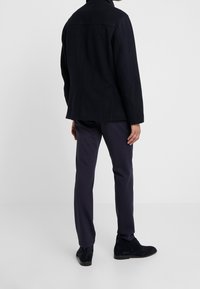 Hombre con una chaqueta negra, pantalones azul marino oscuros y zapatos de ante negros, de pie sobre un fondo blanco sin costuras, de espaldas.