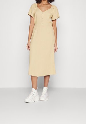 Mujer con un vestido midi beige de mangas cortas abullonadas y botones, combinado con zapatillas blancas altas y gruesas, de pie sobre un fondo liso.