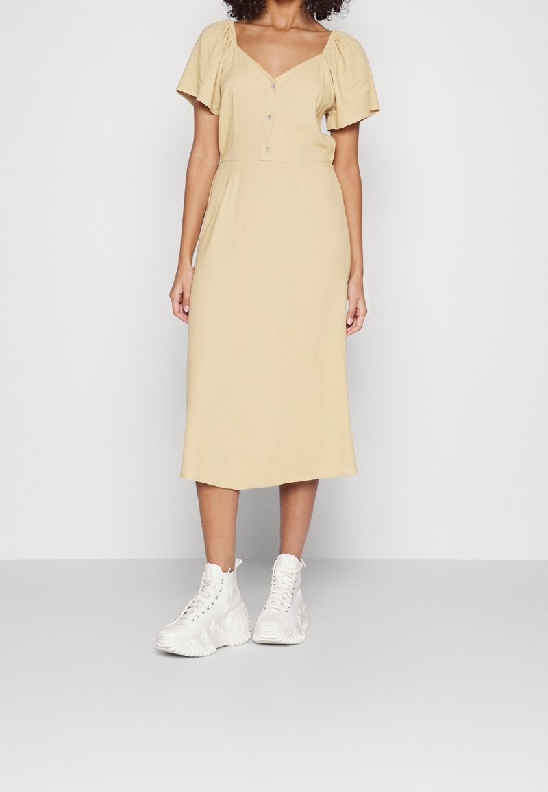 Femme portant une robe midi beige avec des manches courtes bouffantes et des boutons, associée à des baskets blanches montantes à semelle épaisse, debout sur un fond uni.