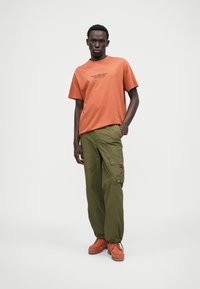 Tricou din bumbac portocalie cu text negru, asortat cu pantaloni cargo verzi cu buzunare laterale și o patch cu logo. Pantofii maro completează lookul.