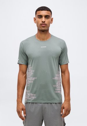 Joven con camiseta de manga corta Salomon verde claro con patrones geométricos blancos en los laterales y pantalones cortos grises, de pie sobre fondo liso.