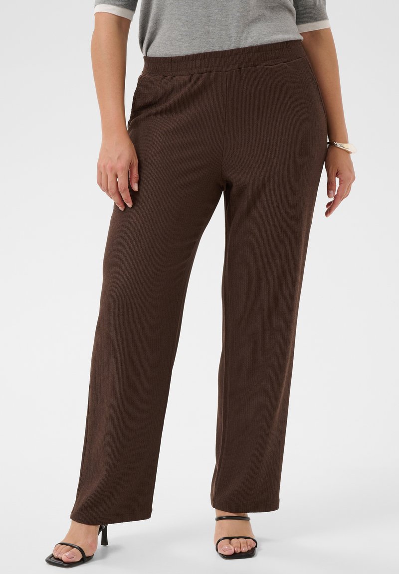 Femme portant un pantalon marron côtelé, ample, avec une taille élastique et des sandales noires à talons ouverts, debout devant un fond uni.