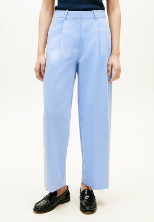 Mujer lleva pantalones de pierna ancha de tiro alto en azul claro y mocasines negros, de pie contra un fondo blanco liso.