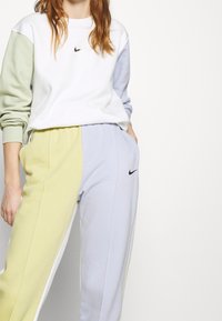 Sweatshirt Nike blanc avec des manches vertes et bleues, associé à un pantalon de jogging avec des panneaux jaunes et bleus, arborant des logos noirs.