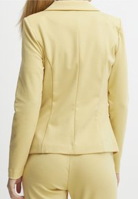 Blazer ajusté jaune en tissu doux, avec un col cranté et des coutures structurées pour un look sur mesure. Vue de dos affichée.