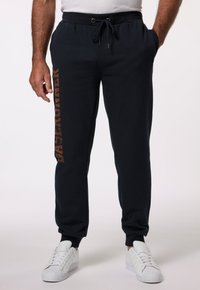 Marina blå sweatpants med dragsko i midjan, sidofickor och tryckt "BASE RUNNER"-design i brunt längs vänster ben. Kombineras med vita sneakers.
