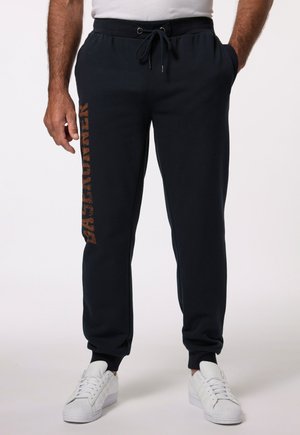 Marineblauwe sweatpants met een elastische tailleband, zijzakken en een geprinte "BASE RUNNER" design in bruin langs het linkerbeen. Gecombineerd met witte sneakers.
