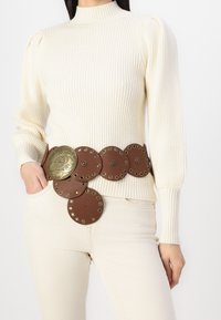 Brązowy skórzany pasek z dużymi okrągłymi dyskami i ozdobnym mosiężnym środkiem, wyposażony w metalowe nity oraz misternie wygrawerowany wzór.