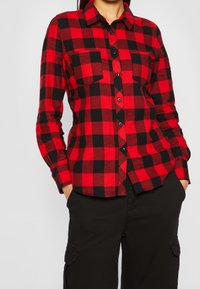 Camicia di flanella a quadri rossi e neri con bottoni e taschino sul petto; presenta maniche lunghe e un orlo arrotondato. La texture sembra morbida.