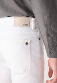 Bijele traperice od denima s kožnom oznakom brenda "BOSS" na leđima, s malim logotipom na džepu i kontrastnom sivom majicom.