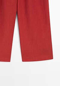 Onderste deel van rode wijde broek met zichtbare stofstructuur, tegen een effen witte achtergrond.