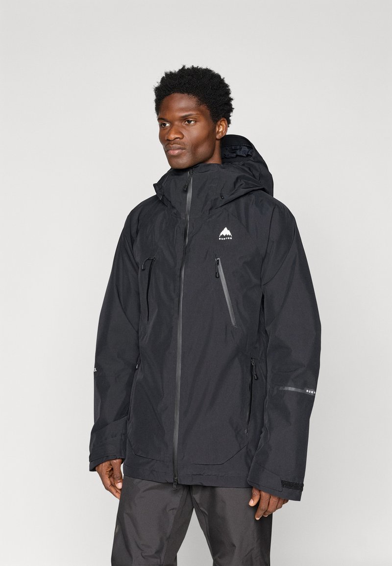 Casaco impermeável preto com capuz, duas pockets frontais com fecho, e um logótipo no lado superior esquerdo do peito. Apresenta punhos elásticos e um fecho robusto.