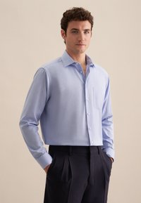 Camicia azzurra chiara a maniche lunghe con bottoni, tessuto morbido, colletto a punte, tasca singola sul petto, abbinata a pantaloni scuri.