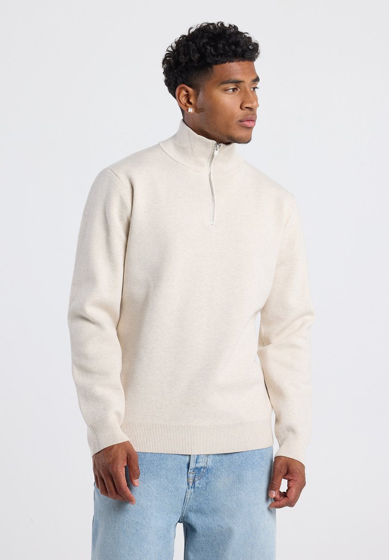Junger Mann mit kurzen lockigen Haaren, bekleidet mit einem beigen Pullover mit Reißverschluss und hellblauen Jeans, schaut nach rechts vor einem schlichten Hintergrund.