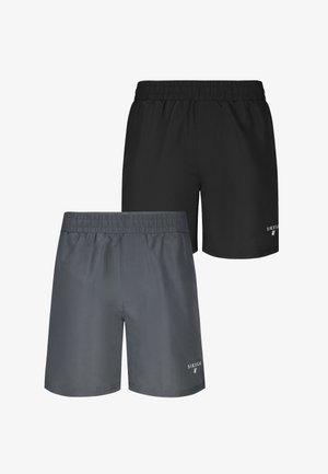 Homme musclé avec de nombreux tatouages, dont un grand visage sur le torse, portant un short foncé, debout pieds nus devant un fond uni.