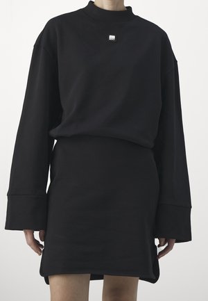 Persoon die een oversized zwarte top met lange mouwen en een bijpassende zwarte rok draagt met een kleine vierkante speld op de bovenborst.