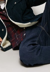 Corduroy jas in donkerblauw met een crèmekleurige fleece voering, gedragen over een bordeauxrode en witte geruite blouse. Donkere spijkerbroek met lichte stiknaden.