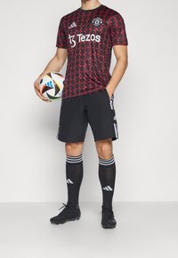 Mies, jolla on Manchester Unitedin jalkapallopaita, mustat shortsit ja sukat, pitelee moniväristä jalkapalloa ja seisoo yksiväristä taustaa vasten.