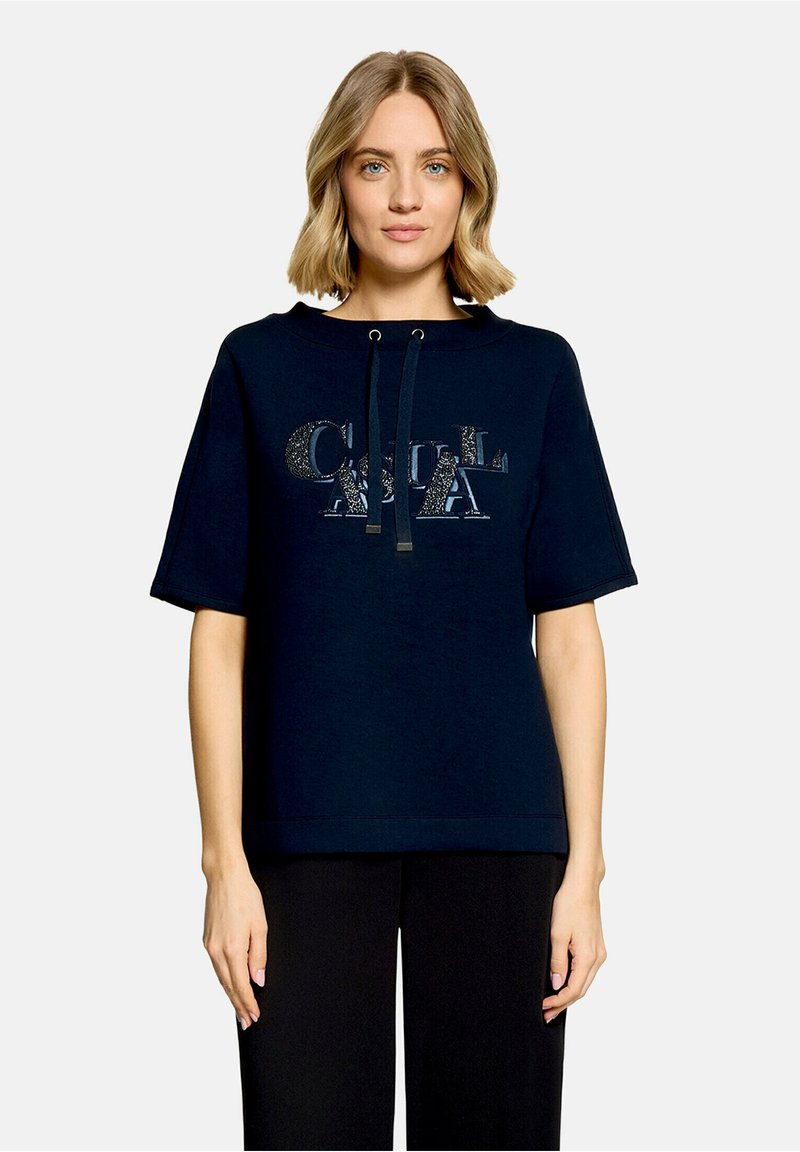 Femme aux cheveux blonds courts portant un sweat à capuche bleu marine foncé à manches courtes avec un logo brillant et un pantalon noir sur un fond uni.