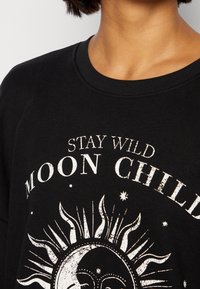 Sudadera negra con cuello redondo que presenta el texto blanco "STAY WILD MOON CHILD" y un diseño gráfico de sol. El material parece suave y texturizado.