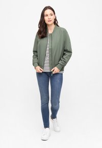 Olivgrön bomberjacka med rund halsringning, matchad med en randig skjorta och blå skinny jeans. Vita sneakers fulländar outfiten.