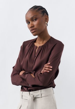 Γυναίκα με πλεξούδες, που φοράει βυσσινί μπλούζα και μπεζ παντελόνι ψηλόμεσο με ζώνη κορδόνι, με σταυρωμένα τα χέρια, μπροστά σε ανοιχτό φόντο.