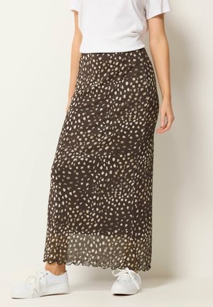 Kobieta w czarnej spódnicy midi w beżowe nieregularne plamy, białej koszulce i białych trampkach, stojąca na tle jednolitego jasnego tła.