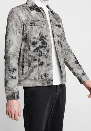 Grijs-zwarte tie-dye denimjas met knoopsluiting, gedragen over een witte coltrui en gecombineerd met zwarte broek, getoond van midden tors tot aan de pols.