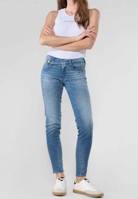 Mujer de pie con los brazos cruzados, vestida con jeans ajustados de color azul claro, camiseta sin mangas blanca y zapatillas blancas con suelas beige.