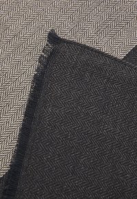 Textiel met een visgraatpatroon in beige en crème kleuren, met een marineblauwe franjes aan één kant, die verschillende texturen toont.