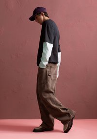 Schwarz-weiße Langarmbluse, die unter einem schwarzen T-Shirt getragen wird, kombiniert mit braunen weiten Hosen und braunen Schuhen vor einem rosa Hintergrund.