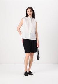 Hvit ermeløs bluse med strukturmønster og knappelukking foran, paret med svarte shorts. Modellen holder en liten svart håndveske.