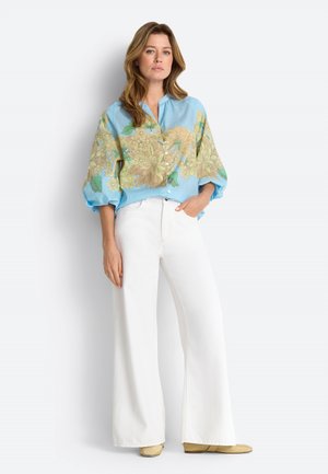 Woman wearing a blue floral button-up shirt with puffed sleeves and white wide-leg pants, standing with one hand in pocket.