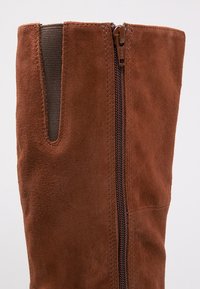 Botte haute jusqu'au genou en suède marron avec fermeture éclair verticale et panneau latéral élastique pour ajustement, présentée sur un fond clair uni.